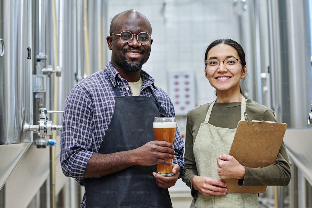 Portrait of couple of brewers smiling at camera while degustating beer together in brewery