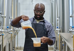 아프리카계 미국인 엔지니어가 갓 양조한 맥주의 품질을 확인하고 맥주를 한 주전자에서 다른 주전자로 붓습니다