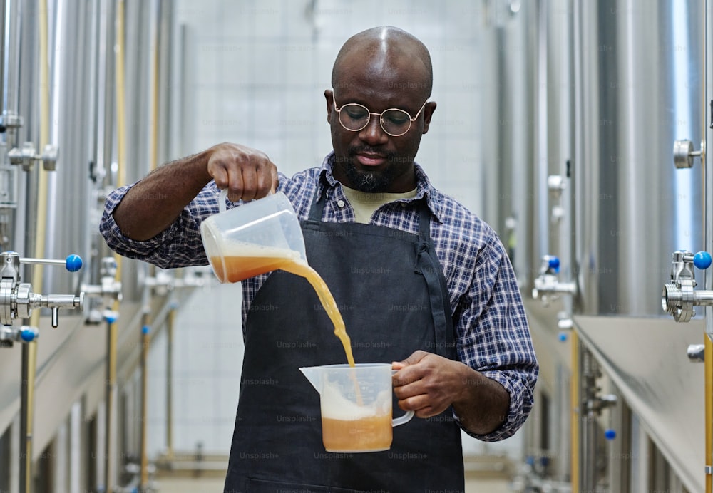African American engineer checking the quality of fresh brewed beer, he pouring the beer from one jug to other