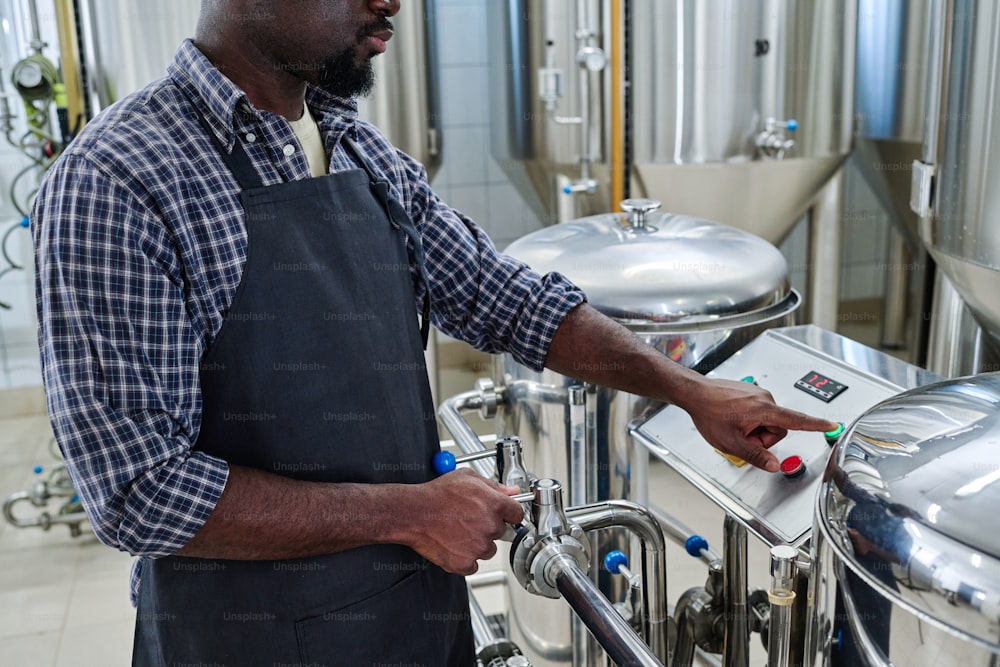 Close-up of African American worker in apron pushing the button to turn on the equipment for brewed beer