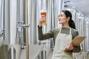 Specialist with document looking at glass of fresh beer to check the quality during her work at brewing plant
