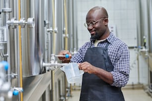 Trabajador afroamericano vertiendo cerveza fresca para probarla después de prepararla mientras está de pie en el taller