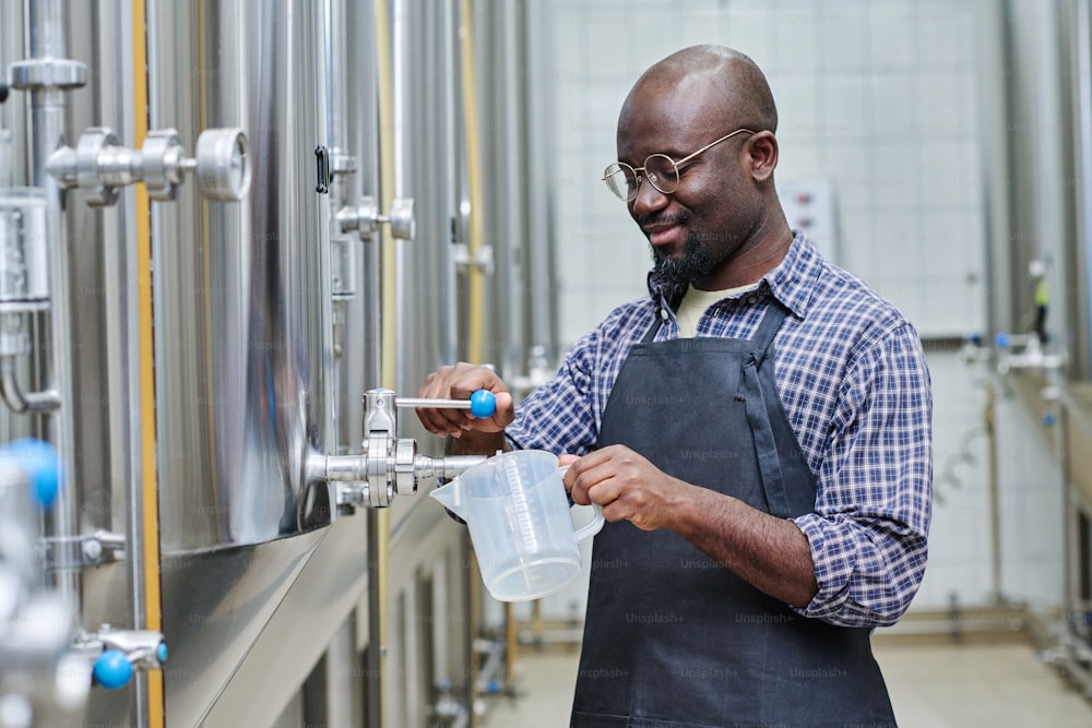 African American worker pouring fresh beer to taste it after brewing while standing in workshop