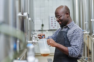 Trabajador afroamericano de una fábrica vierte cerveza elaborada en un recipiente para su degustación después del procesamiento