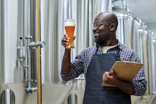 Smiling African American worker holding glass of fresh beer to check the quality after brewing while standing in workshop with tanks