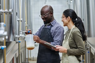 Pareja multiétnica de ingenieros que vierten cerveza fresca del barril para probarla durante su trabajo en la fábrica de cerveza