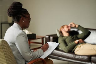 African American psychologist making notes in card during her work with patient while he lying on sofa in background