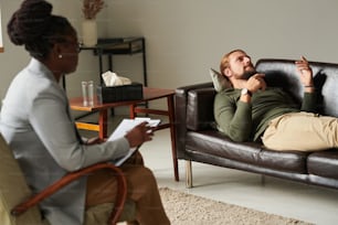 Man lying on sofa and discussing his problems with psychotherapist while she making notes during consultation at office