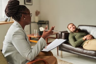 Afroamerikanischer Psychologe mit Dokument, der auf dem Sessel sitzt und mit dem Patienten im Büro spricht