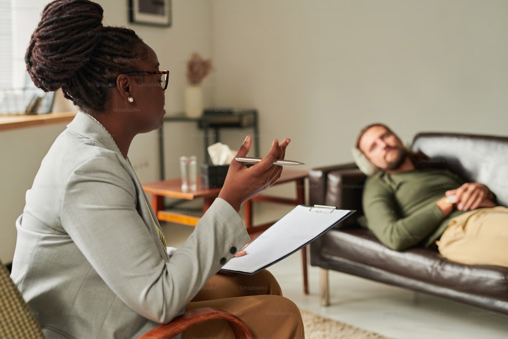 African American psychologist with document sitting on armchair and talking to patient at office