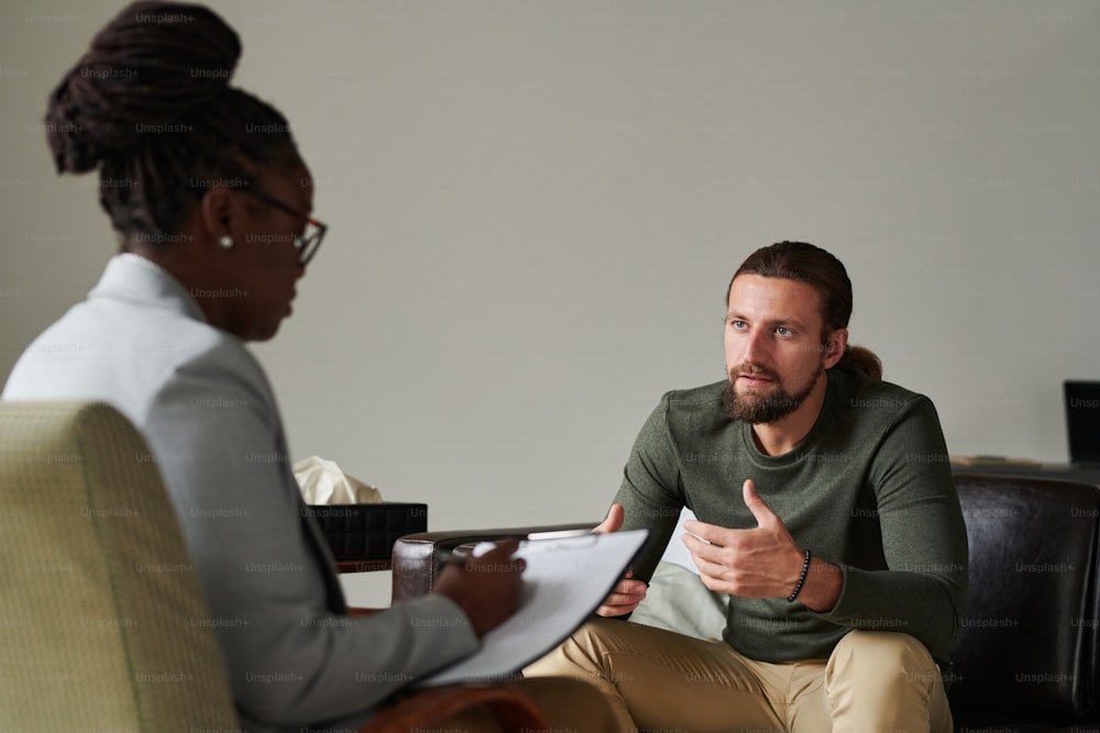 Young man having consultation with psychotherapist, he talking to her while she making notes