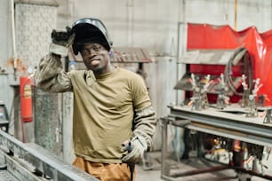 Retrato de un joven soldador africano con máscara sonriendo a la cámara mientras trabaja con productos metálicos en una fábrica