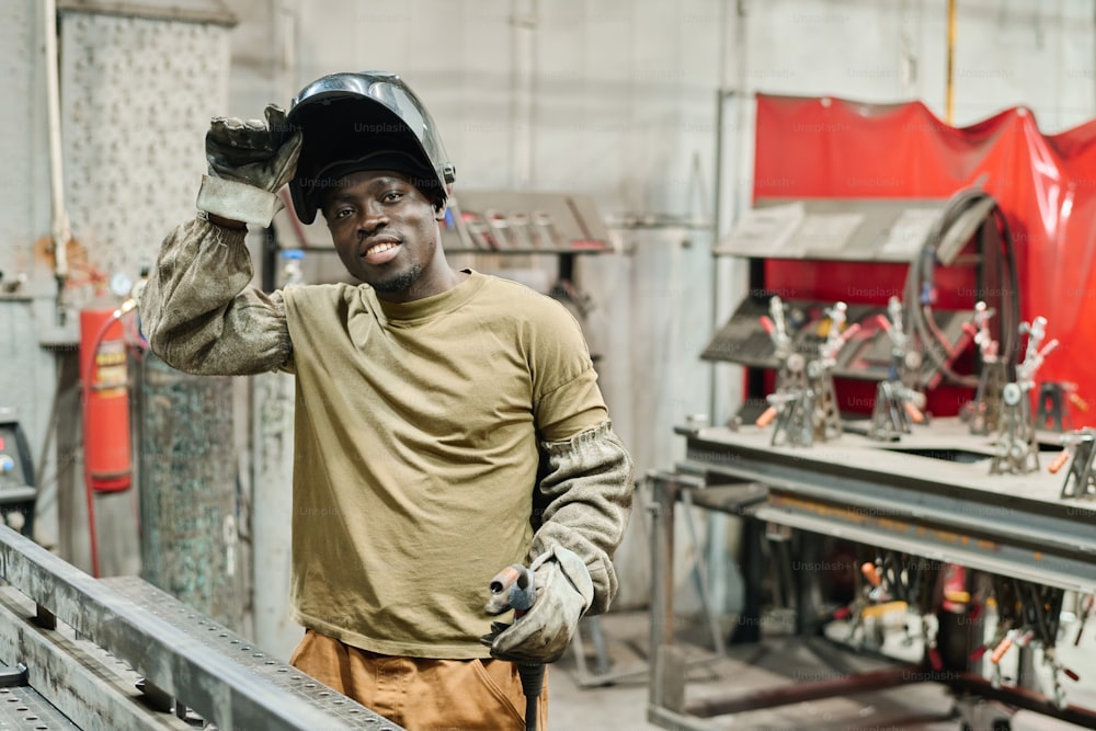 Retrato de un joven soldador africano con máscara sonriendo a la cámara mientras trabaja con productos metálicos en una fábrica