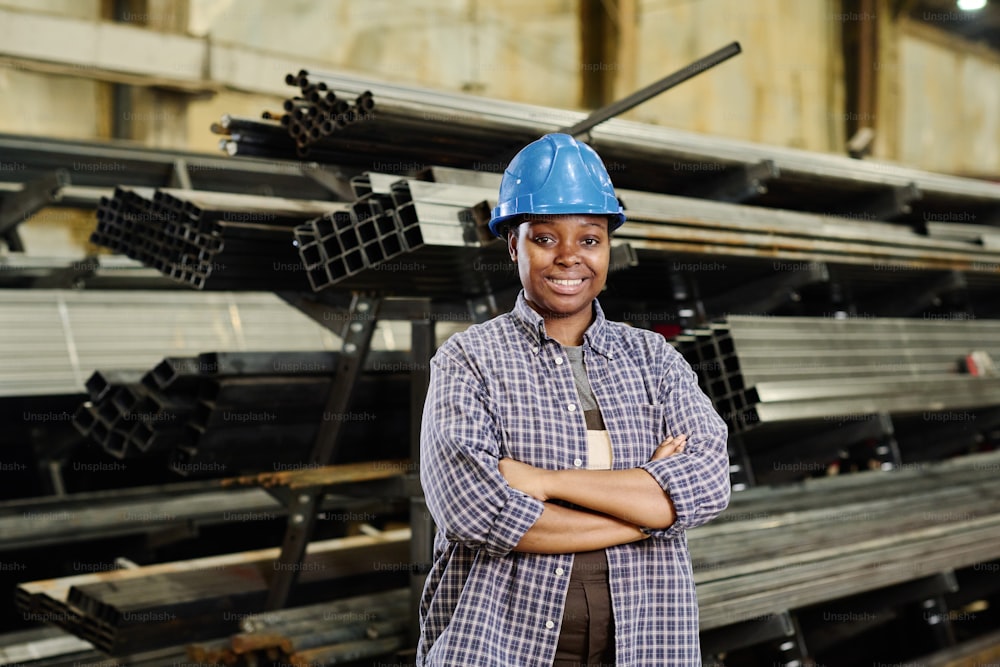 Portrait d’une métallurgiste africaine portant un casque de sécurité souriant à la caméra debout, les bras croisés contre des piles métalliques