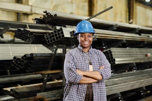 Portrait of African female metalworker in hardhat smiling at camera standing with her arms crossed against metal piles