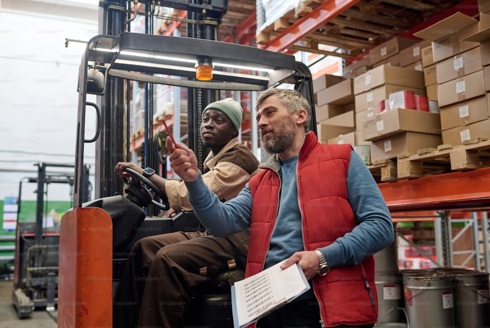 Manager with checklist giving instruction to forklift worker during their work at warehouse