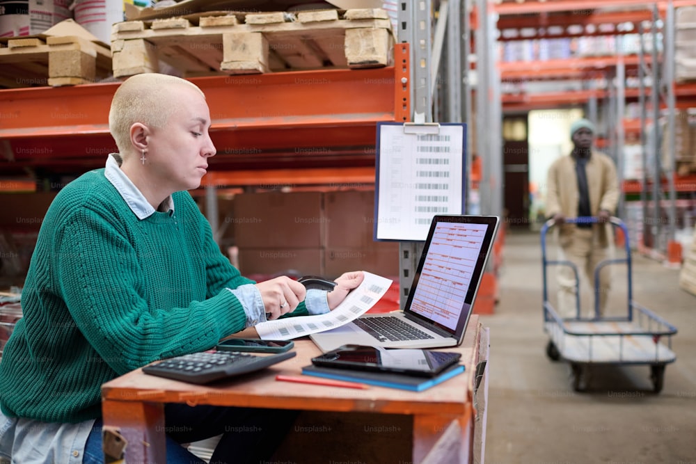 Serious woman sitting at table with laptop using scanner to scan barcodes of received parcels during her work at warehouse