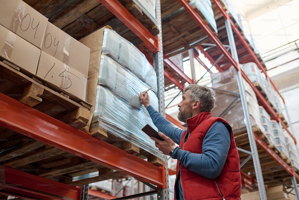 Mature worker using tablet pc and noting packages on shelves at warehouse before shipping