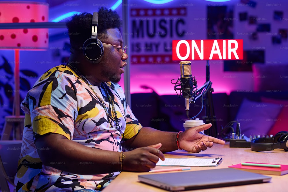 Vista lateral do belo jovem afro-americano usando fones de ouvido sentado na mesa de gravação de podcast ou transmissão em estúdio em casa