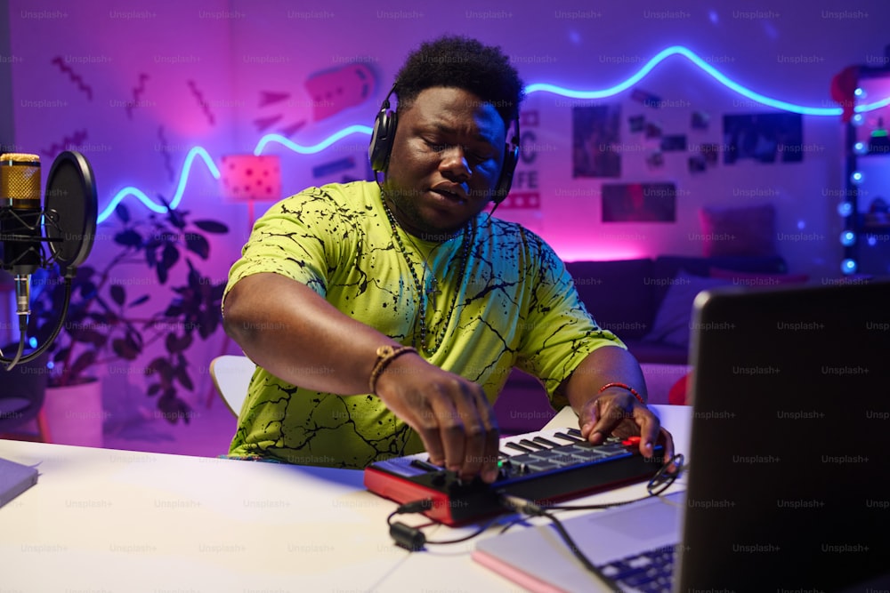 Porträt eines jungen schwarzen Musikers der Generation Z, der Kopfhörer trägt, zu Hause am Schreibtisch sitzt und mit Synthesizer und Laptop an der Audiospur arbeitet