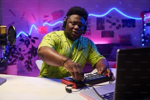 Retrato del joven músico negro de la generación Z con auriculares sentado en el escritorio de su casa trabajando en una pista de audio usando sintetizador y computadora portátil