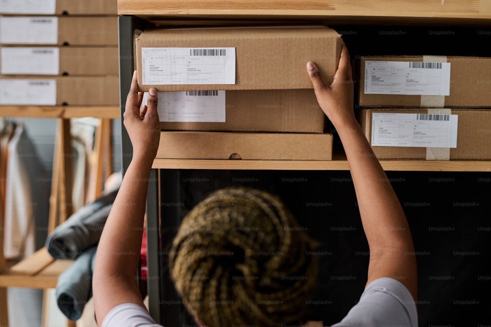 Bras d’une jeune femme noire triant des boîtes en carton emballées avec des marchandises commandées par des clients en ligne et en prenant une sur une étagère en bois