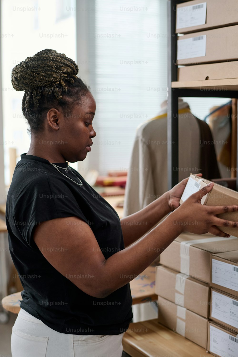 Young female worker of online shop warehouse sorting parcels and checking addresses of receivers while standing by shelf