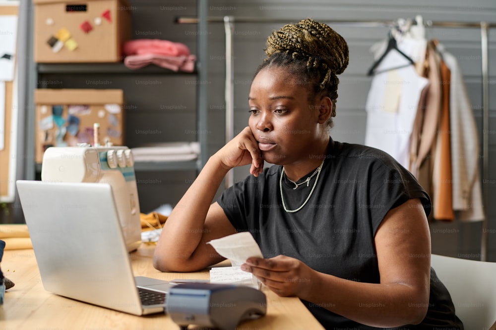 Young serious black woman with receit looking at laptop screen by workplace while checking payment of online clients