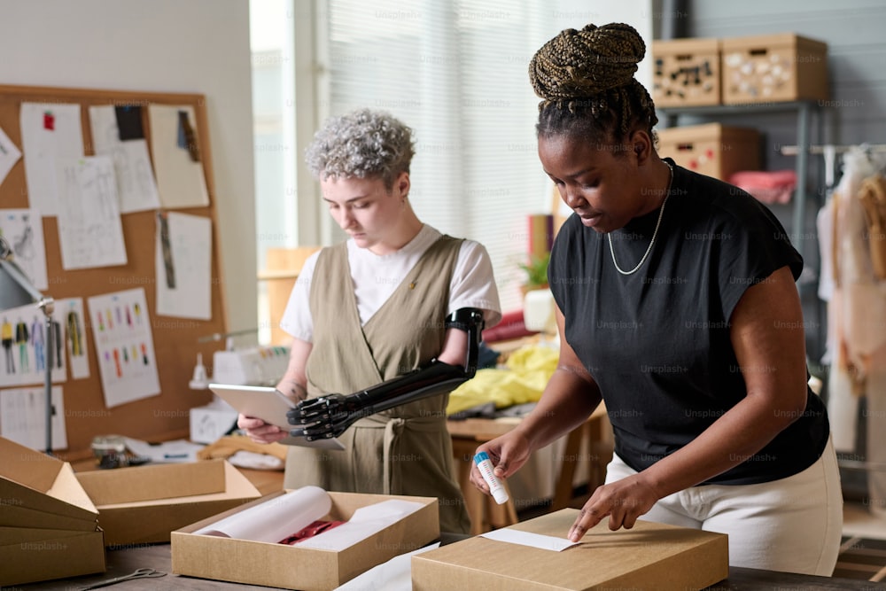 Black woman sticking small paper with receiver address on packed box with goods while her colleague with disability using tablet