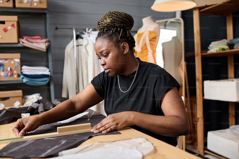 Young serious African American female tailor making textile pattern for new clothes while drawing line with chalk close to wooden ruler