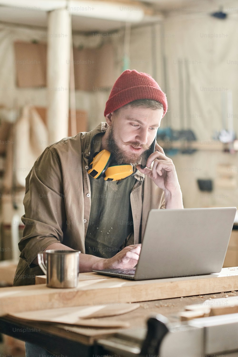 Modern male carpenter with beard on face talking on phone while typing on laptop in woodworking workshop