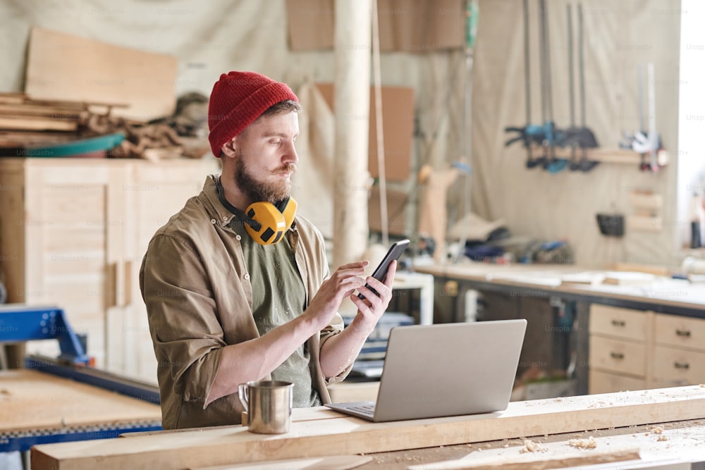 Moderner Holzarbeiter, der mit Laptop und einer Tasse Tee an der Werkbank steht und etwas auf dem Smartphone schreibt