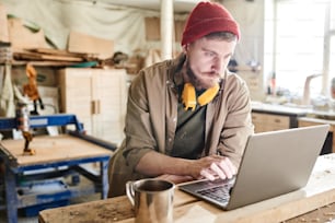 Junger bärtiger Zimmermann mit Gehörschutz über dem Hals surft in der Kaffeepause auf dem Laptop im Internet