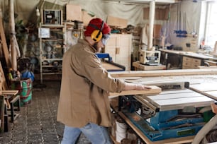 Carpintero profesional con gafas de seguridad y auriculares protectores cortando tablón de madera con sierra de mesa