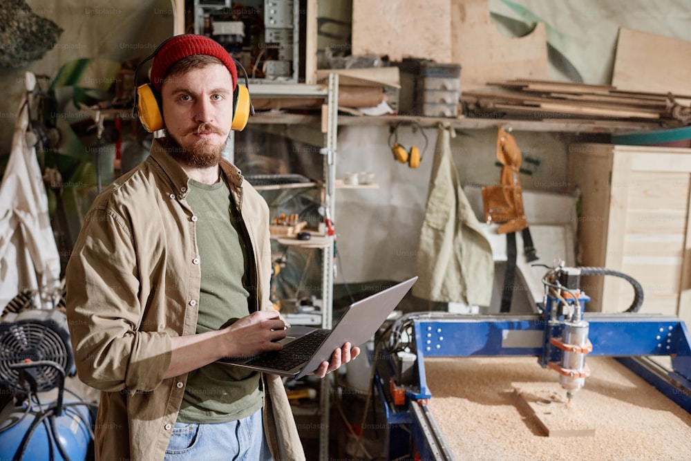 Horizontal medium portrait of young Caucasian man working in carpentry workshop holding laptop looking at camera