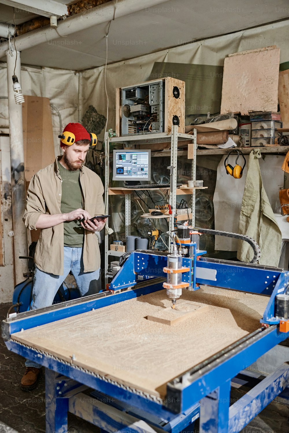 Vertical long shot of young Caucasian man using tablet while operating CNC milling machine in woodworking workshop