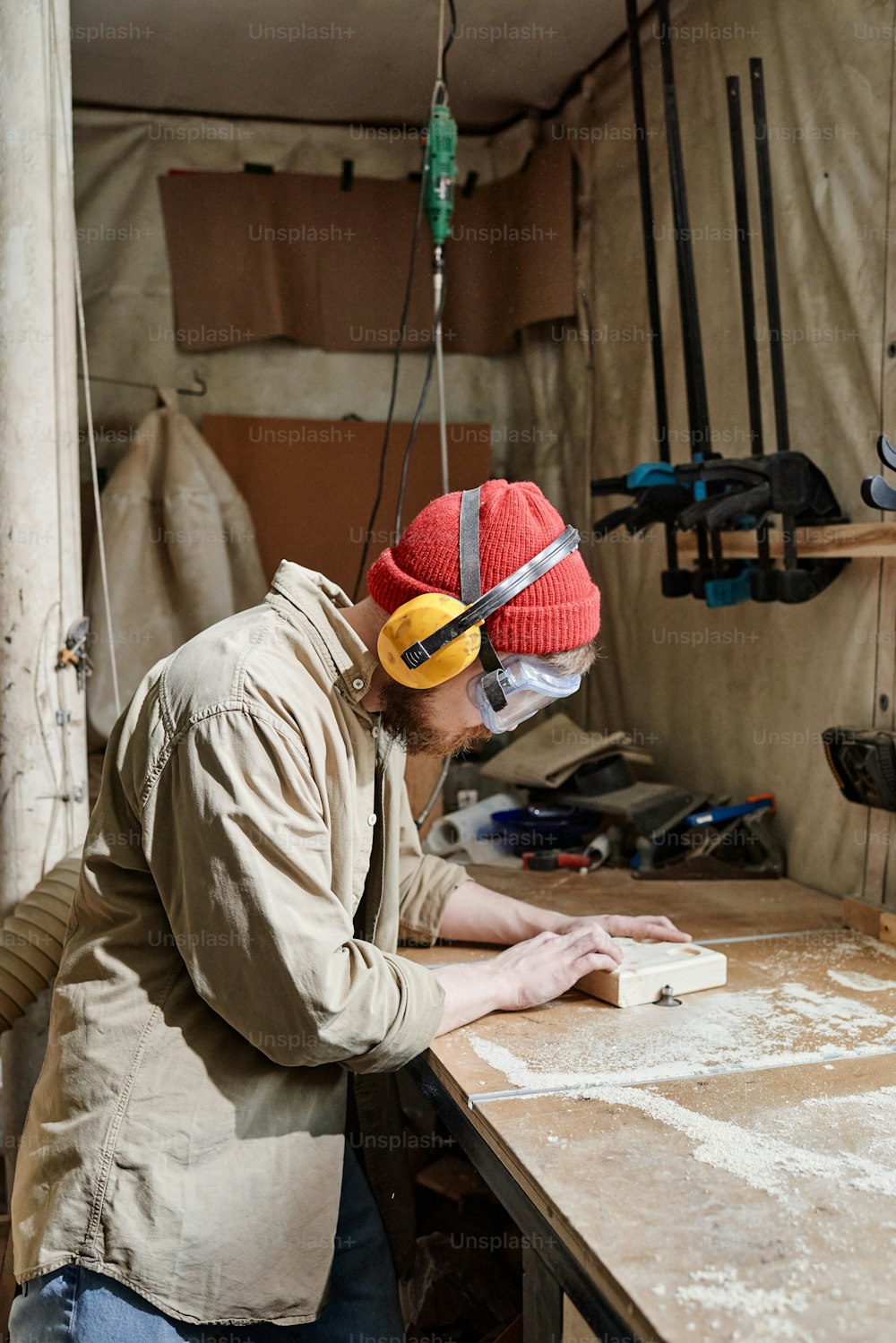 Vertical shot of young man wearing safety glasses and earmuffs shaping wooden detail using benchtop router table in woodworking workshop