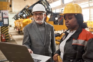 Ingénieur principal en casque de travail utilisant un ordinateur portable pour présenter de nouvelles machines aux travailleurs qui regardent un moniteur debout près