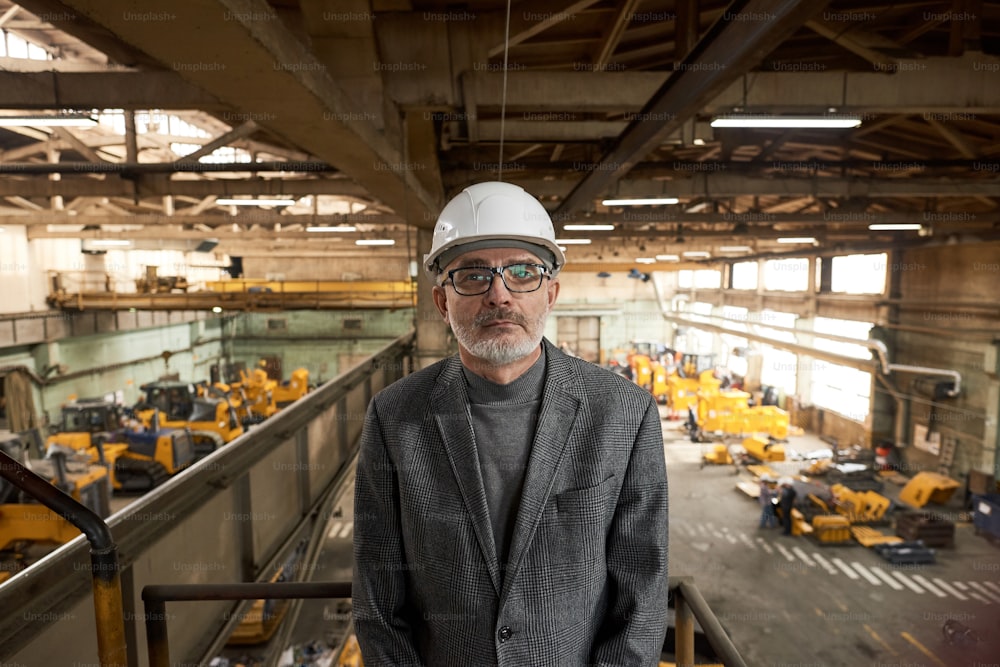 Portrait of senior architect in work helmet looking at camera while standing on balcony in plant with production equipment in the background