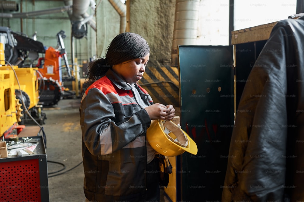 Femme africaine en uniforme debout près de son placard et enlevant le casque de travail après son travail à l’usine
