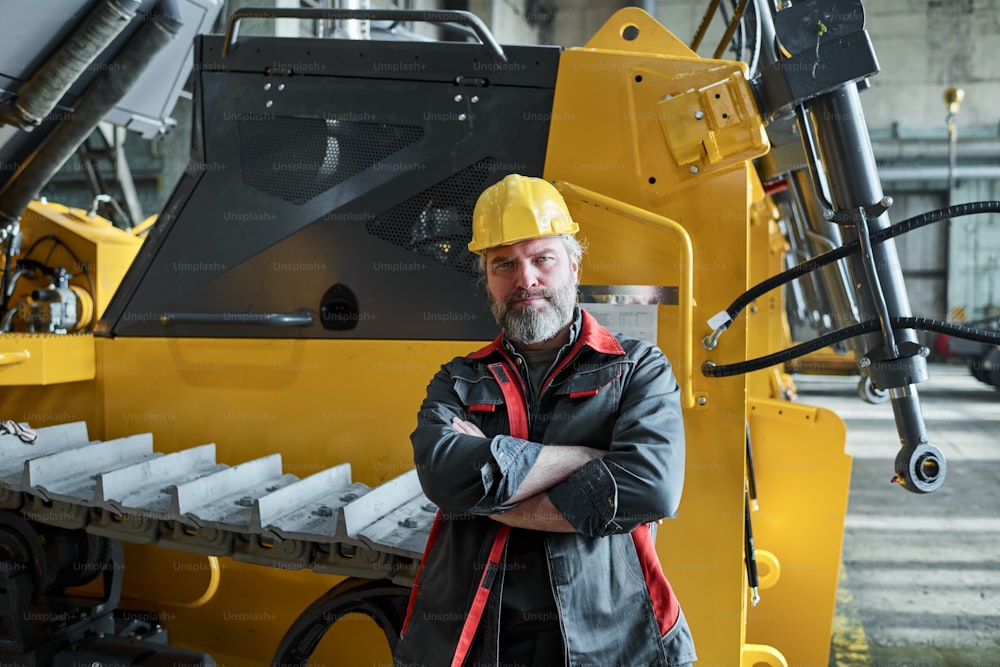 Portrait of mature bearded worker in uniform standing with arms crossed against the big machine and looking at camera
