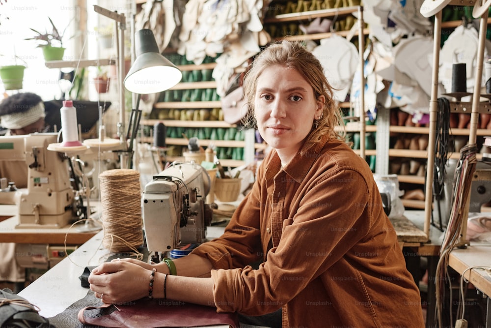 Portrait of young dressmaker looking at camera while sitting at the table with sewing machine thread and pieces of fabric in the workshop