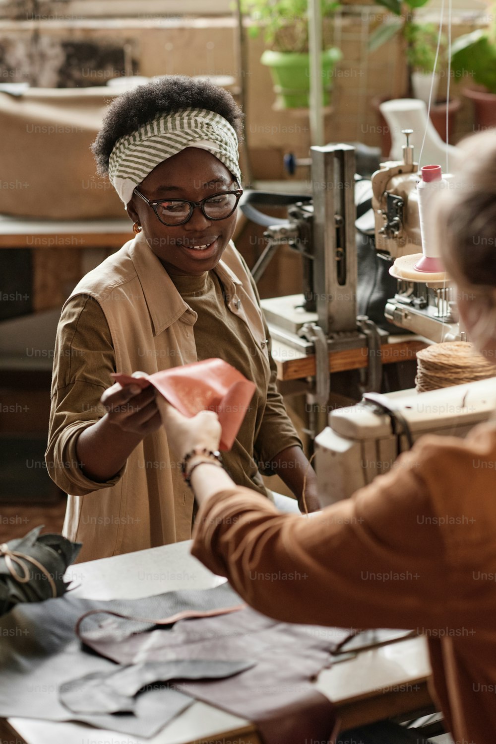 African young woman discussing the piece of leather together with her colleague while they sewing on sewing machines in the workshop