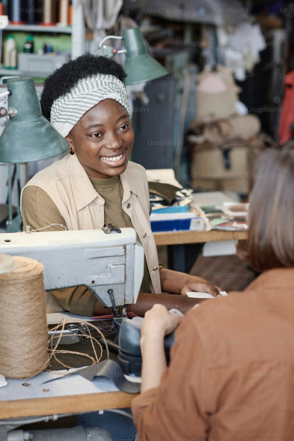 African young tailor smiling to her colleague while they talking during their sewing work in the workshop
