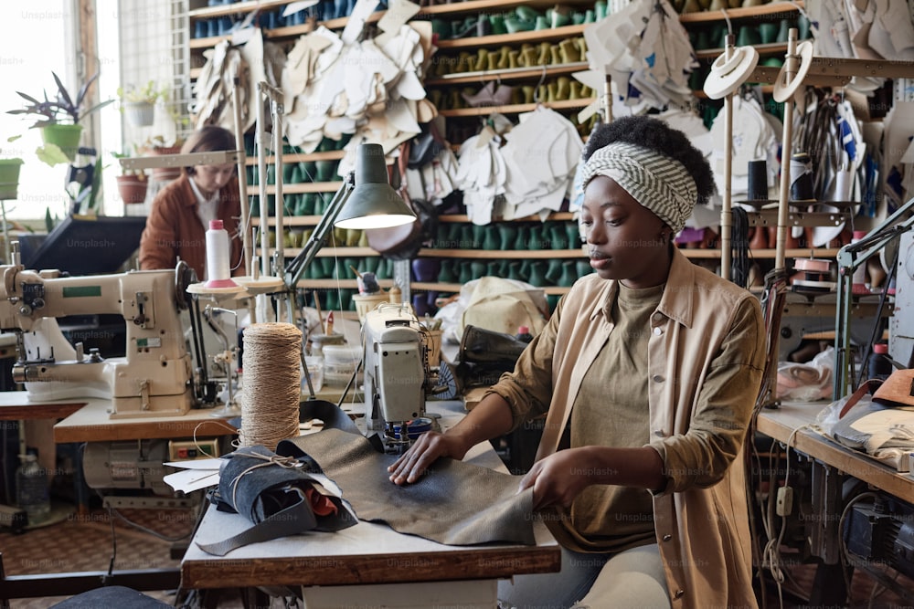 African tailor sitting at her workplace with sewing machine and sewing products in factory