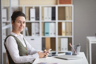 Portrait of young businesswoman looking at camera while sitting at workplace and using her mobile phone at office