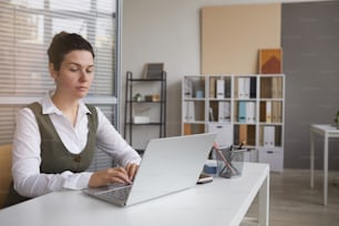 Oficinista seria concentrada en su trabajo, se sienta a la mesa y escribe en una computadora portátil en la oficina