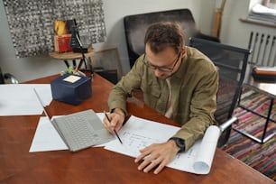 Young architect in eyeglasses sitting at his workplace and examining the blueprint of new building