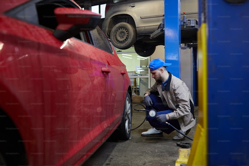 Horizontal side view shot of professional repairman sitting in front of car wheel using tire pressure gage