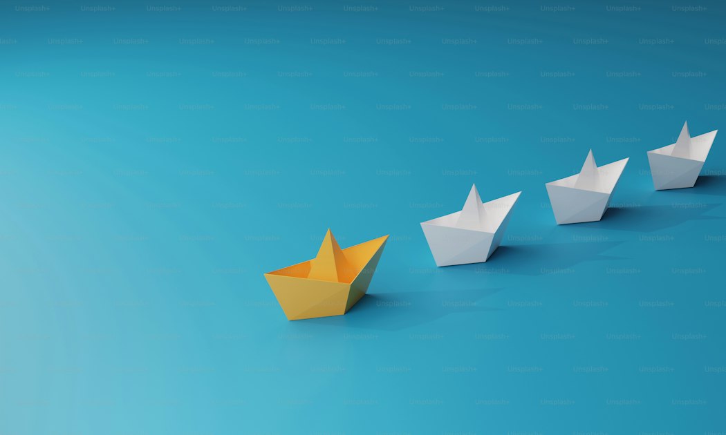 A yellow paper boat leading a group of white paper boats on a blue background